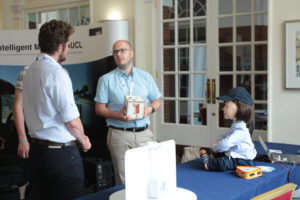 Two men in blue shirts discuss how to interact with the robot child figure seated on the table