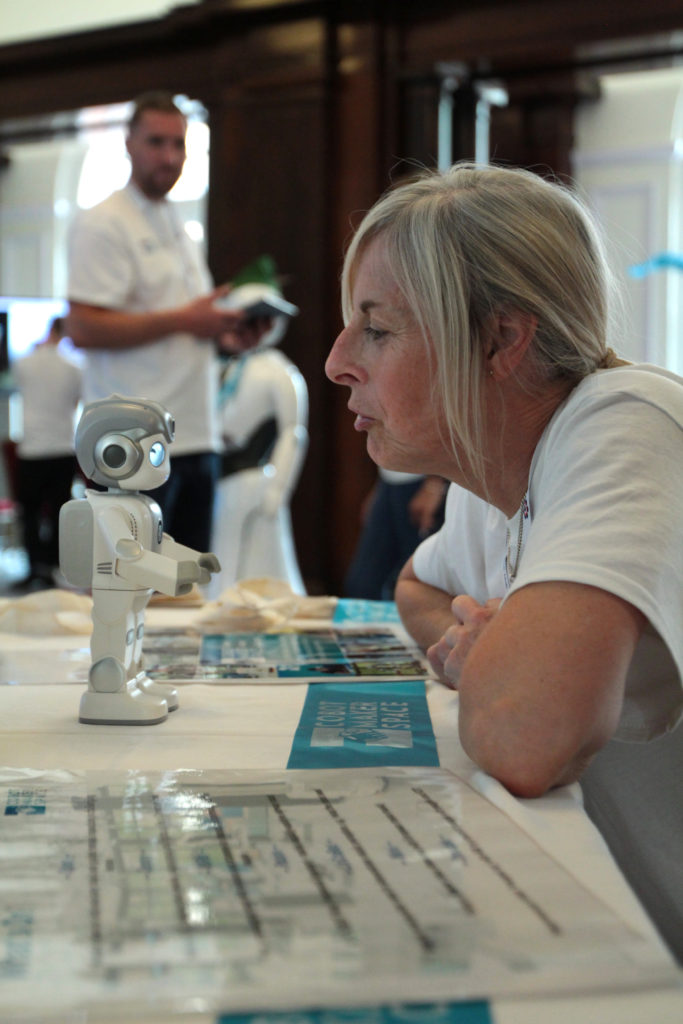 Hazel Sayers interacts with small robot, man in white stands in the background